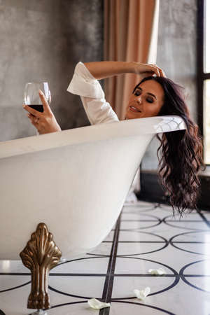 Happy fashion lady lying in retro bath with goblet of red wine relaxing at luxury apartmentの写真素材
