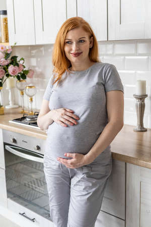 Happy pregnant woman posing with coffee tea mug in modern minimalistic nordic kitchen at homeの写真素材