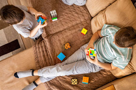 Top view two teenage and baby brothers assembling multicolored Rubiks cubes sitting on couchのeditorial素材