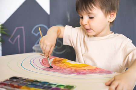 Confident cute male kid art drawing multicolored rainbow arch use paints and brush on tableの写真素材