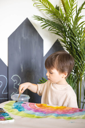 Confident cute male kid art drawing multicolored rainbow arch use paints and brush on tableの写真素材