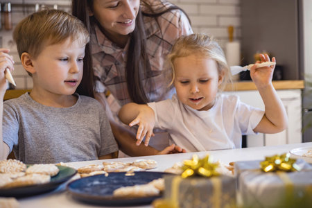 Mom and children decorate Christmas gingerbread at home. A boy and a girl paint with cornets with sugar icing on cookies. New Years decor, branches of a Christmas tree.の写真素材
