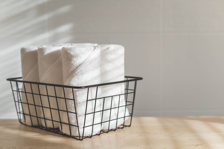 Fresh white towels rolled fluffy fabric spa textile stack in metallic basket wooden sunny tabletopの写真素材
