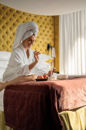 Relaxed happy woman sitting on luxury bed with champagne glass and breakfastの写真素材