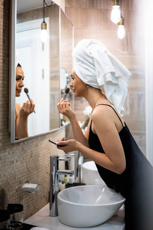 Happy brunette woman applying makeup powder blush foundation use brush in front mirror at bathroomの写真素材