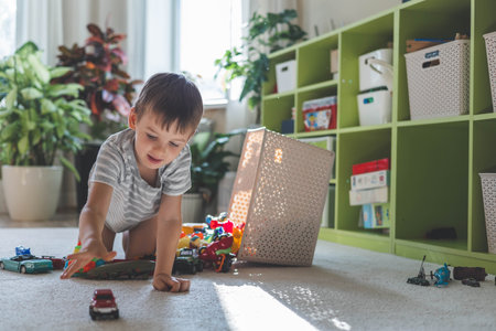 A cheerful European boy plays with cars on the carpet in his room.