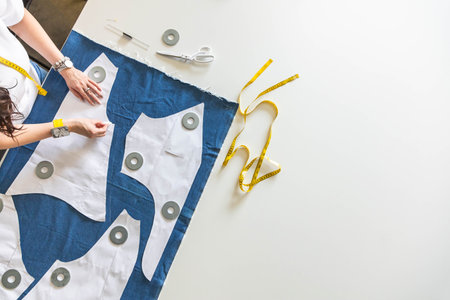 A woman dressmaker lays out pattern details on denim and cuts clothes. Top view.