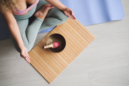 Woman in lotus position practicing yoga singing meditation bowl on wooden board top view closeupの写真素材