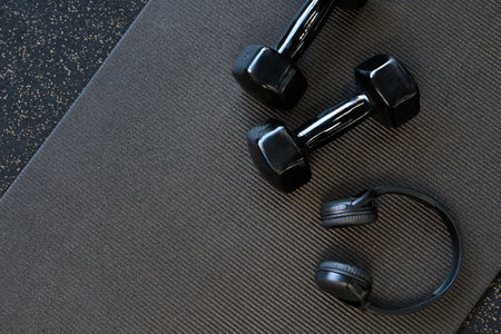Headphones and black dumbbells lie on a black yoga mat. Flat lay. Space for text.