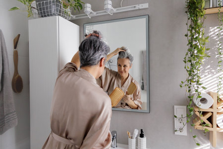 An elderly woman in her ages, a pensioner in silk pajamas, examines her gray hair holding a comb.の写真素材
