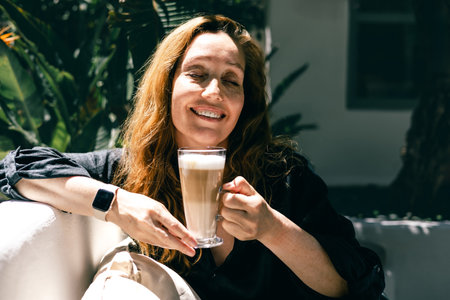 Smiling Woman Enjoying Latte in Sunlit Garden with Relaxed Mood and Natural Vibesの写真素材