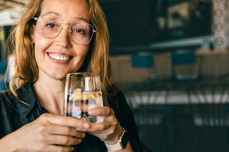 Happy Woman Holding Glass of Water with Lemon, Smiling in Cozy Cafe, Healthy Lifestyleの写真素材