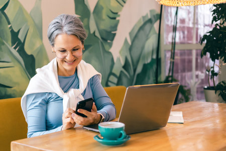 Happy senior freelancer woman using smartphone while working from cozy cafe with laptop