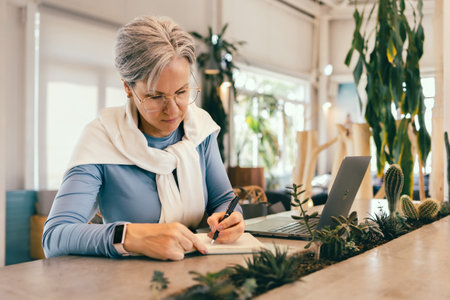 Focused senior woman with gray hair writing in notebook while working on laptop in cozy modern cafeの写真素材