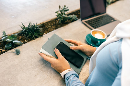 Close-up of woman holding smartphone and notebook in cafe with coffee and laptopの写真素材