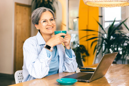 Happy senior freelance woman enjoying coffee break while working remotely in bright modern cafe