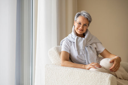 A mature, elderly, aged, beautiful and well-groomed woman sits in a white chair near the windowの写真素材