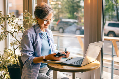 Senior businesswoman writing notes from phone at cafe table with laptop, smiling, working remotelyの写真素材