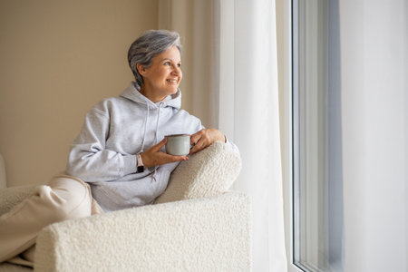 Senior woman relaxing at home with cup of coffee near window in cozy hoodie and soft armchair..の写真素材