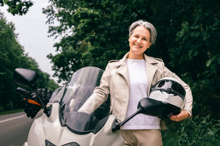 Smiling gray-haired senior woman in beige outfit holding motorcycle helmet on scenic country roadの写真素材