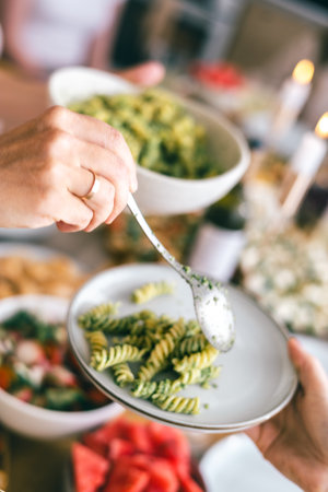 Close-up of hands serving green pesto pasta with spoon on white plate during festive summer mealの写真素材