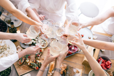 Friends clinking glasses of white wine over festive lunch table with appetizers, cheese, bruschettaの写真素材