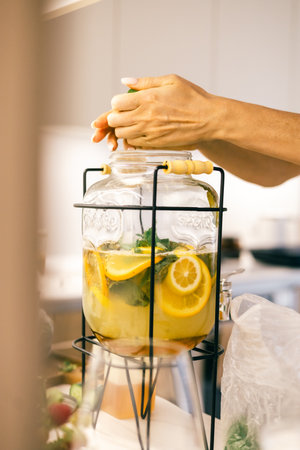 Close up of hands preparing citrus infused water in glass dispenser with lemons, mint in kitchen.の写真素材