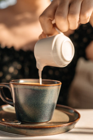 Pouring cream into ceramic cup of hot coffee in sunlight close up of morning breakfast moment