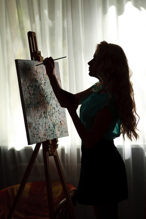 Silhouette woman artist draws paint picture on easel, backlight portrait indoorの写真素材