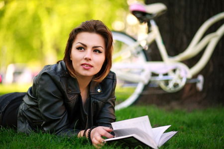 Young woman with bicycle reading a book in a city park on the grass
