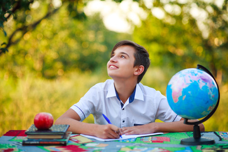 Dreamy schoolboy pupil studying at a table in a garden outdoors with a smile looking up. School...