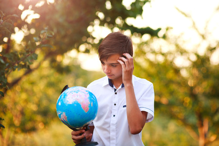 Thoughtful boy teenager holding a globe of planet earth in a summer park on a background of the nature trees at sunset, concept save the planetの写真素材