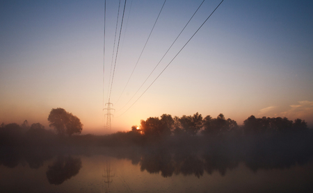 Electrical steel pylon and cable in the haze at sunset in forest above river, energy concept in wild natureの写真素材