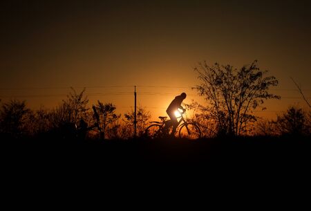 Silhouette of a boy with a bicycle on the background of bright sunset sun in vintage old style, the concept of a carefree childhoodの写真素材