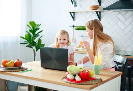 Mother with baby daughter sitting at the table with groceries in the kitchen with a laptopの写真素材
