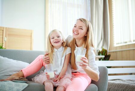 Young mother with child daughter at home on the couch drinking milk and cookies, laughing and talking togetherの写真素材