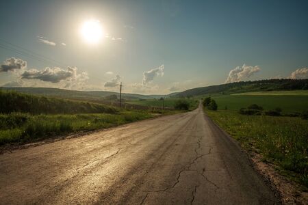Road in the countryside among fields of old cracked asphalt on a background of hills and sky