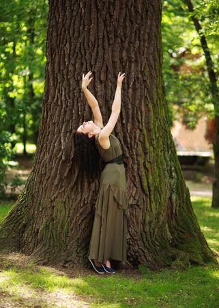 Young woman in a green dress meditatively relaxes near a large tree in a forest park, concept of purity of nature and unity with the environmentの写真素材