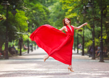 Young woman in a luxury long red dress dancing, jumping in city parkの写真素材