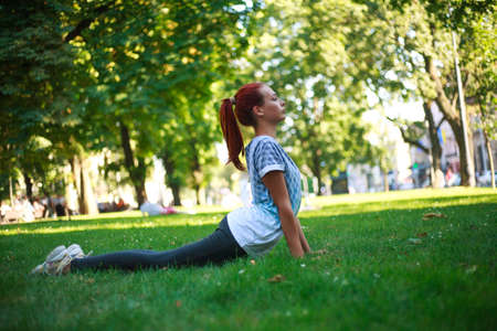 Young woman girl in city park on green grass doing stretching fitness exercises outdoorsの写真素材