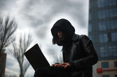 Girl in a black leather coat and a hood with laptop on a city street, concept of a woman hacker and agent working remotely at computerの写真素材