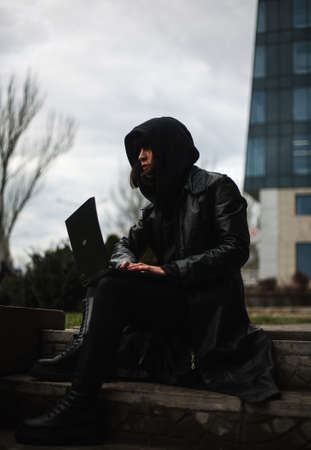 Girl in a black leather coat and a hood with laptop on a city street, concept of a woman hacker and agent working remotely at computerの写真素材