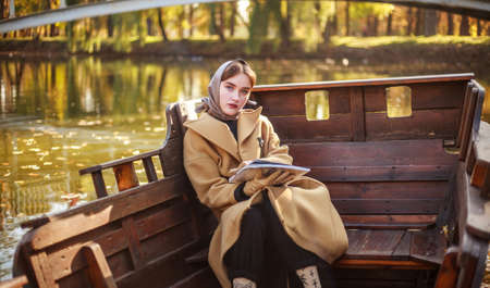 Elegant young lady in a long coat with magazine on a wooden boat on the river in autumn parkの写真素材