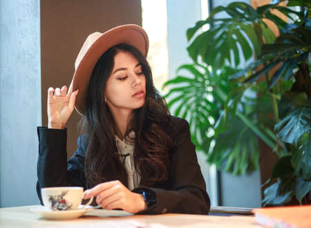 Young Asian business woman in a black jacket and hat in a cafe sits at a table with a cup of tea, on the background of a green plantの写真素材