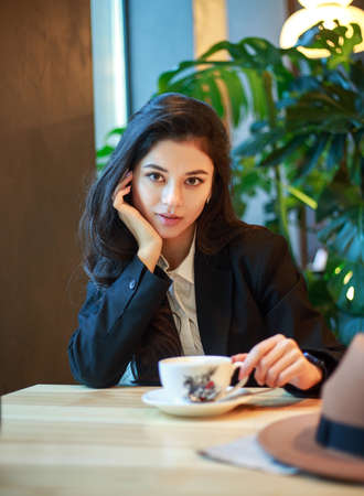 Young Asian business woman in a black jacket and hat in a cafe sits at a table with a cup of tea, on the background of a green plantの写真素材
