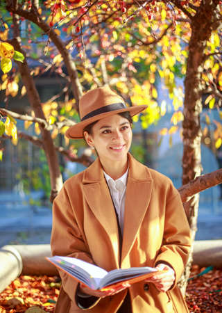 Girl young lady student in a coat and hat in a city park reads a book under a colorful tree, learning outdoorsの写真素材