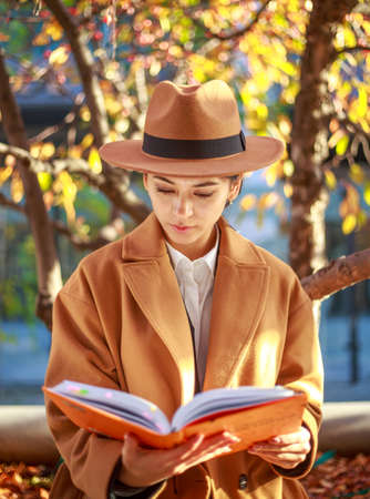 Girl young lady student in a coat and hat in a city park reads a book under a colorful tree,...