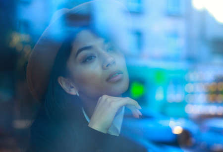 Young asian woman in a classic business style sits pensively peering through the window with reflections of the street and lights with a cup of coffeeの写真素材