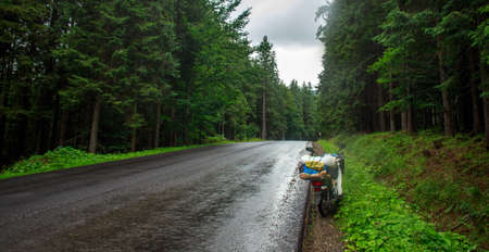 Asphalt empty road in coniferous mountains, traveler's bike moped by the roadside, freedom and journey conceptの写真素材