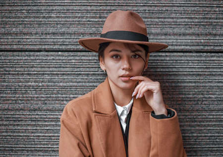 Young business lady in retro style on the background of a stone wall in a brown-beige coat and a classic hat, isolated portrait in natural lightの写真素材